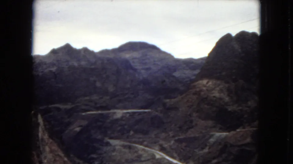 1967 Arizona home-movie frame with dark 8mm frame borders, rocky ridges, a winding road, and canyon terrain in soft faded color.