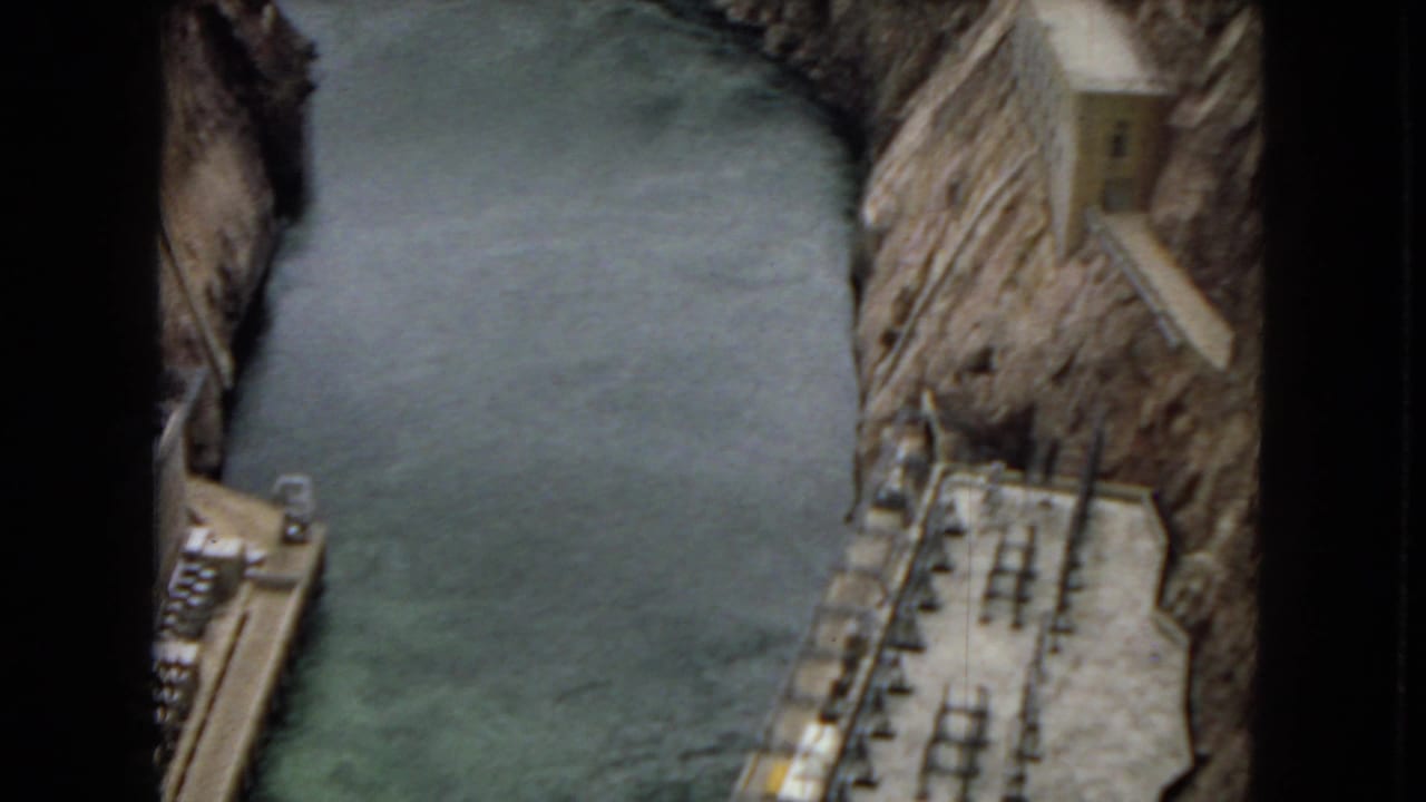1967 Arizona 8mm frame looking down from atop a concrete dam toward green reservoir water, service platforms, and steep rocky canyon walls.