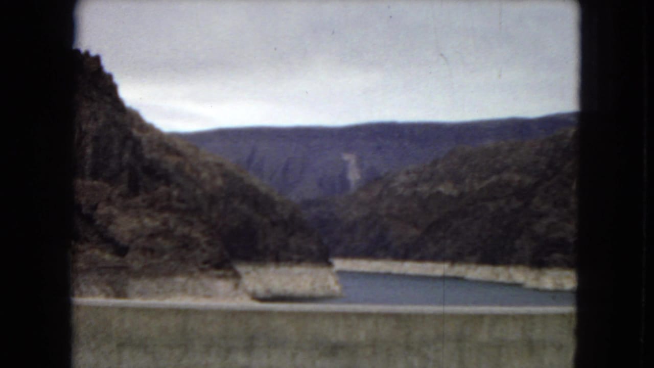 1967 Arizona 8mm frame from a dam overlook showing the reservoir, concrete dam edge, dark canyon slopes, distant mountains, and overcast sky.