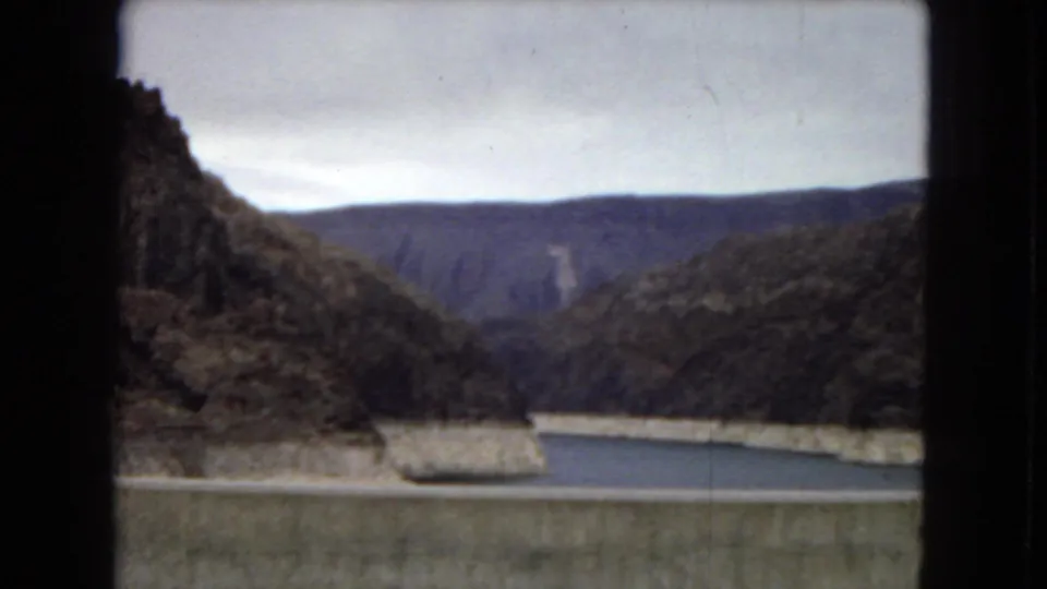 1967 Arizona 8mm frame from a dam overlook showing the reservoir, concrete dam edge, dark canyon slopes, distant mountains, and overcast sky.