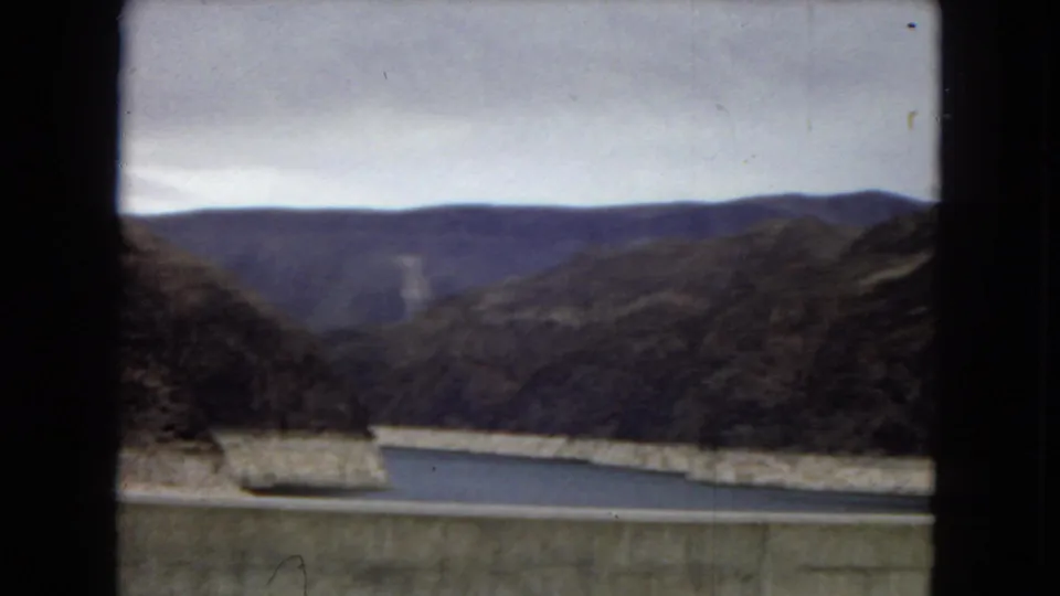 1967 Arizona 8mm frame showing a broad view from the dam across reservoir water, concrete infrastructure, rocky canyon walls, and distant mountain ridges.