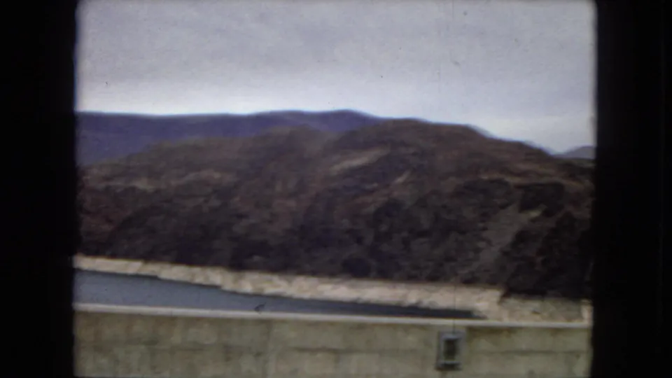 1967 Arizona 8mm frame looking over the concrete dam wall toward reservoir water, rocky desert hills, distant ridgeline, and pale overcast sky.