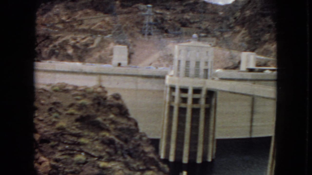 1967 8mm view of Hoover Dam or Boulder Dam, showing the concrete dam face, intake towers, dark water below, and rugged canyon rock behind.