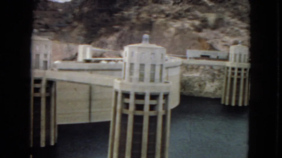 1967 home-movie frame showing an intake tower and dam wall at Hoover Dam or Boulder Dam, with water at the base and rocky desert canyon in the background.