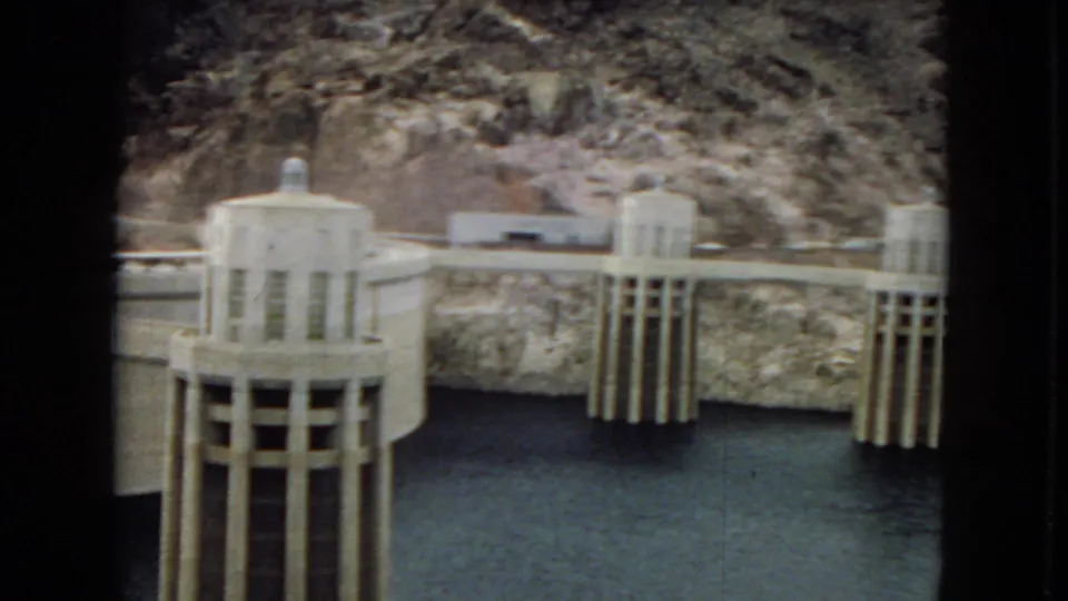 Final 8mm frame of Hoover Dam or Boulder Dam, with twin intake towers, concrete infrastructure, dark reservoir water, and rugged canyon walls visible.