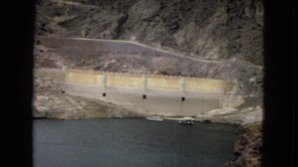 1967 8mm view of a pale concrete dam-side structure below a winding canyon road near Hoover Dam reservoir water.