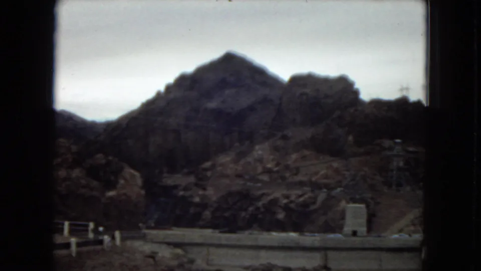 1967 8mm view across Hoover Dam's rocky canyon surroundings, with a dam wall edge and power transmission structures in the distance.