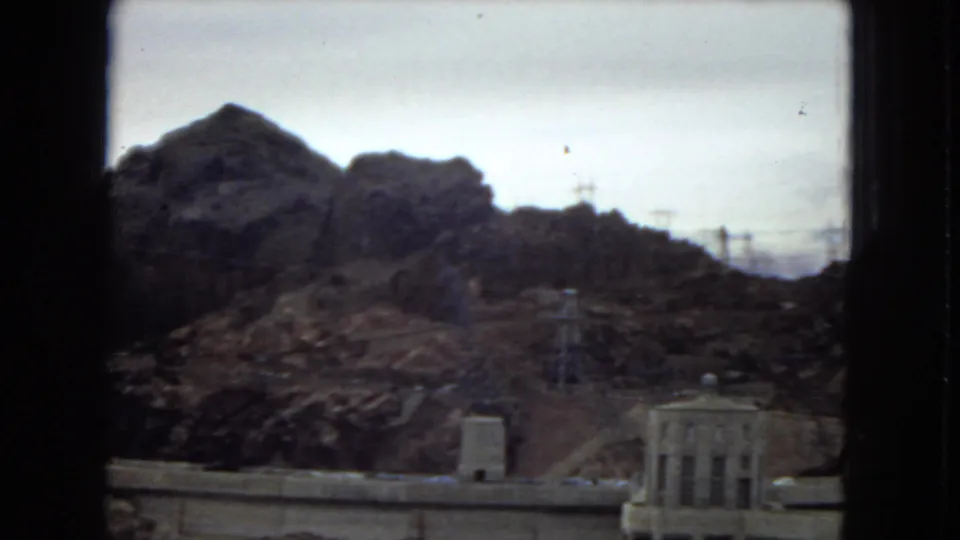 1967 8mm view of Hoover Dam surroundings with canyon slopes, power lines, dam structures, and part of an intake tower at the lower right.