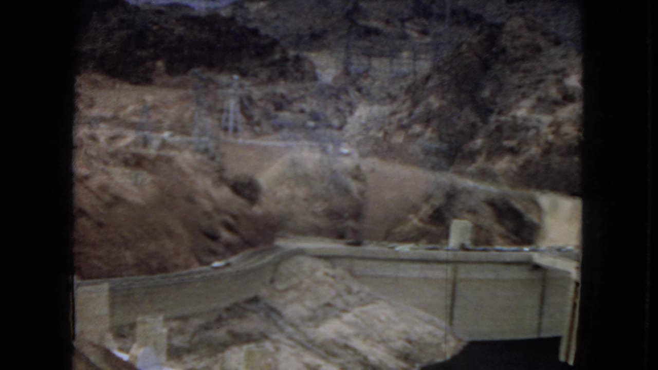 1967 8mm travel frame showing Hoover Dam infrastructure set below arid rocky slopes, with transmission towers visible along the upper ridge.