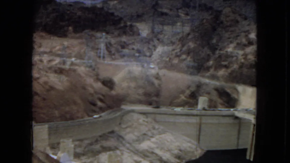 1967 8mm travel frame showing Hoover Dam infrastructure set below arid rocky slopes, with transmission towers visible along the upper ridge.