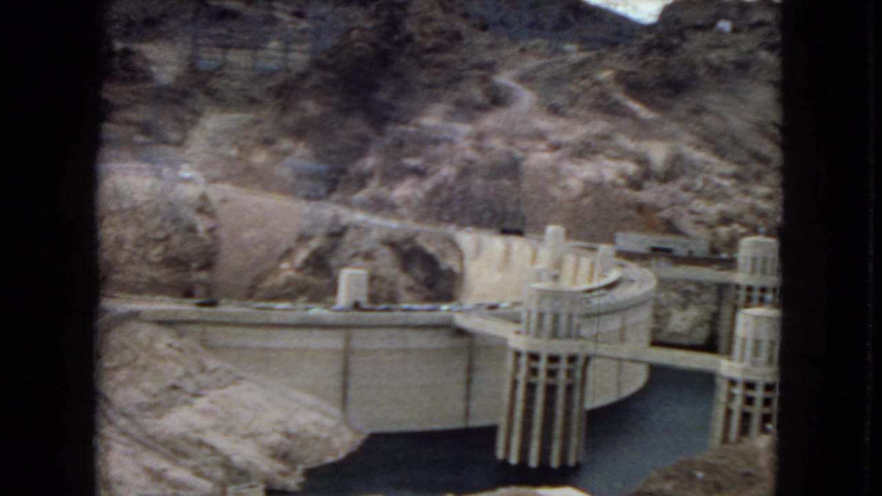 1967 home-movie view of Hoover Dam with cylindrical intake towers, reservoir water, curved concrete works, and dry canyon hillsides.