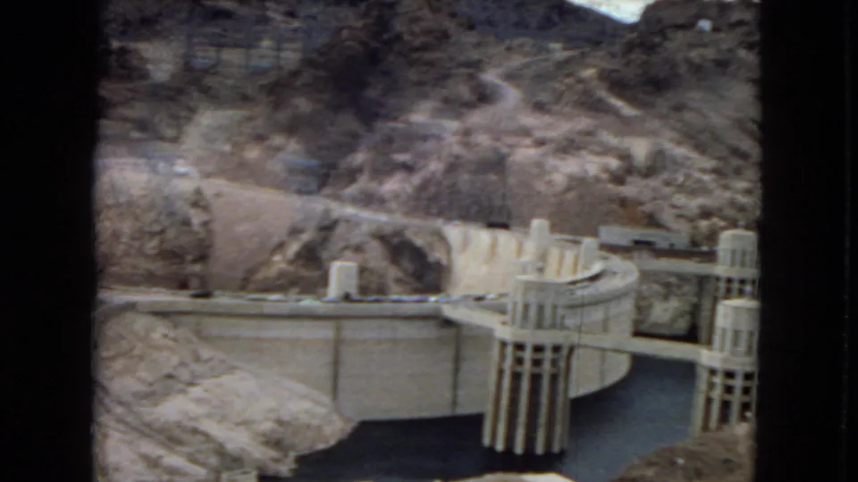 1967 home-movie view of Hoover Dam with cylindrical intake towers, reservoir water, curved concrete works, and dry canyon hillsides.