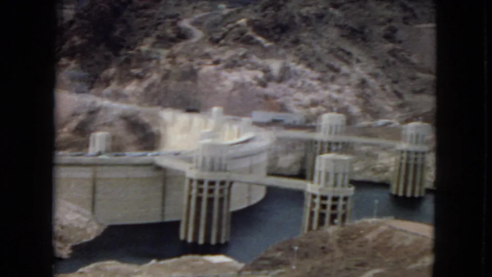 1967 8mm frame of Hoover Dam intake towers and concrete works, with dark reservoir water bordered by rugged arid rock.