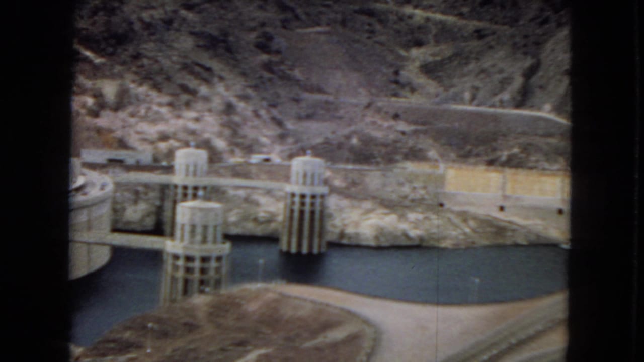 1967 8mm home-movie frame of Hoover Dam with concrete intake towers, reservoir water, a curving road, and rugged desert hills under a cloudy sky.
