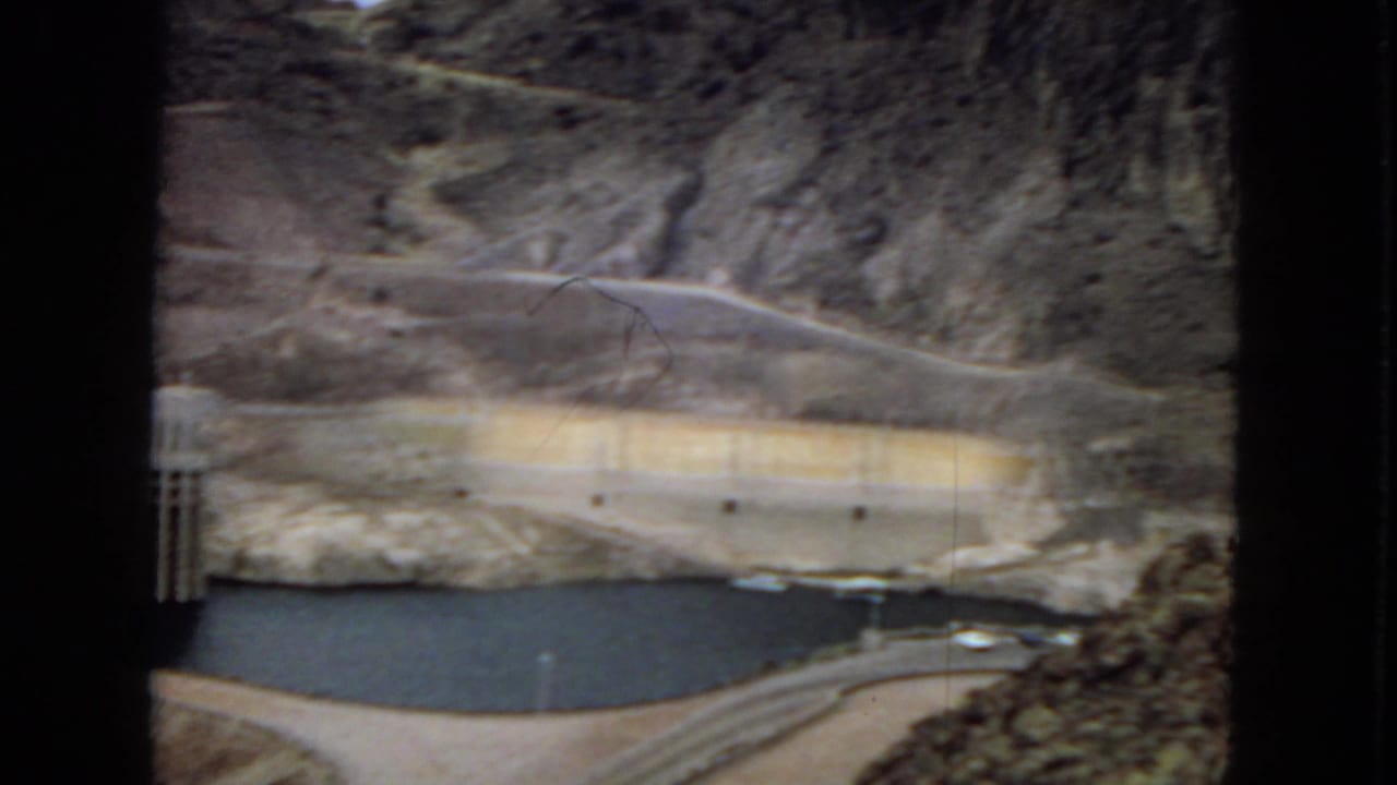 1967 Hoover Dam view showing reservoir water, pale concrete dam surfaces, a roadway along the valley, and steep arid mountainsides.