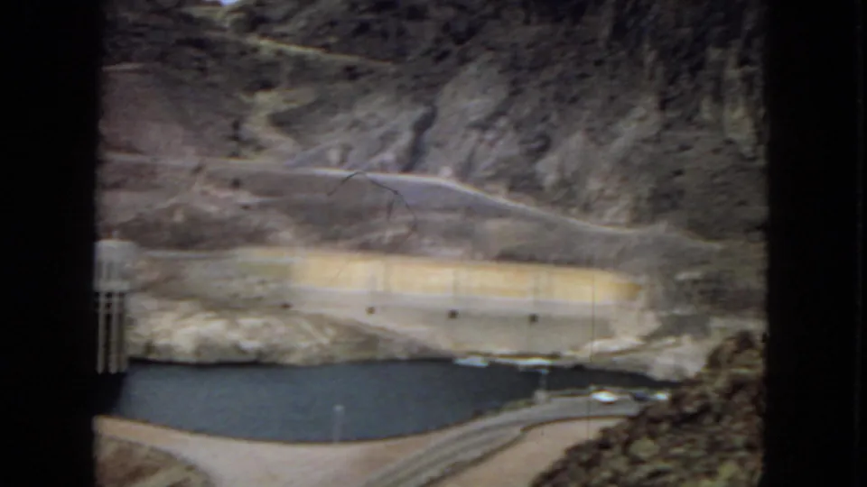 1967 Hoover Dam view showing reservoir water, pale concrete dam surfaces, a roadway along the valley, and steep arid mountainsides.