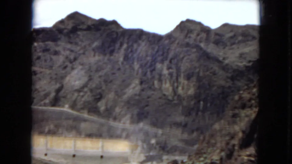 1967 8mm frame looking across Hoover Dam area toward dark rocky mountains, with a dam wall and road visible at the lower edge.