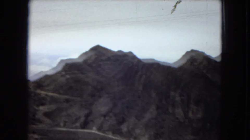 1967 home-movie frame of the Hoover Dam landscape, with rugged mountains, a winding road, pale sky, and dark film borders framing the view.