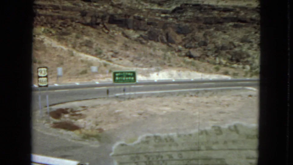 A roadside view along Route 93 shows highway shields, a green directional sign, pavement, and layered desert rock formations in 1967 home-movie footage.
