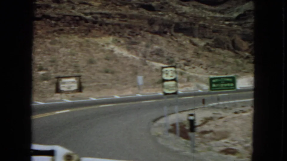 The camera looks across a curving Route 93 roadway with shield signs, a green highway sign, roadside posts, and rocky desert slopes in the background.