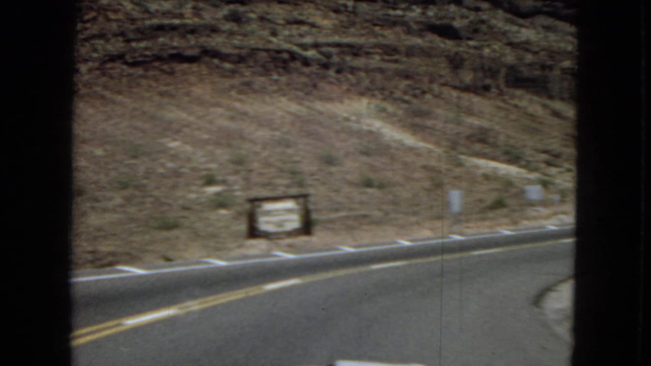 A 1967-supported 8mm view through a car window shows a curving desert road, rocky slope, sparse vegetation, and roadside signs in the American Southwest.