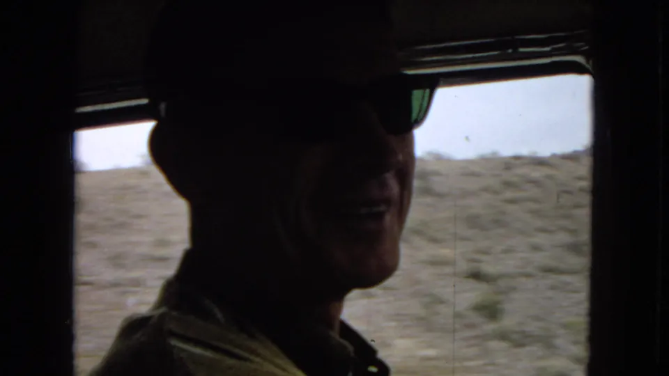 Inside the car, the sunglassed driver appears partly silhouetted as the moving desert landscape blurs through the window behind him.