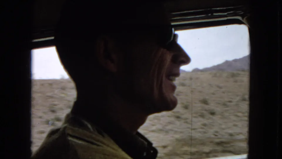 The adult male driver smiles or speaks in profile while desert scrub and low hills pass outside the vehicle window in this 8mm road-trip frame.