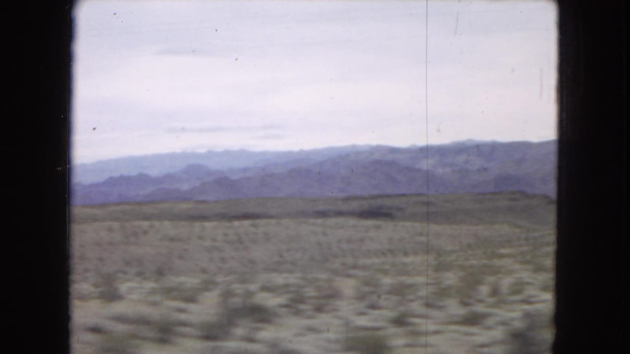 1967 8mm home-movie frame showing arid desert terrain, low scrub, distant mountain ridges, and cloudy sky viewed through a dark vehicle window frame.