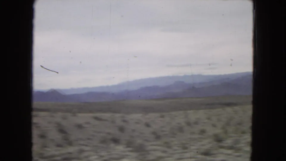 A 1967 road-trip view across pale desert brush toward layered mountains, divided by dark interior vehicle framing and softened by 8mm motion blur.