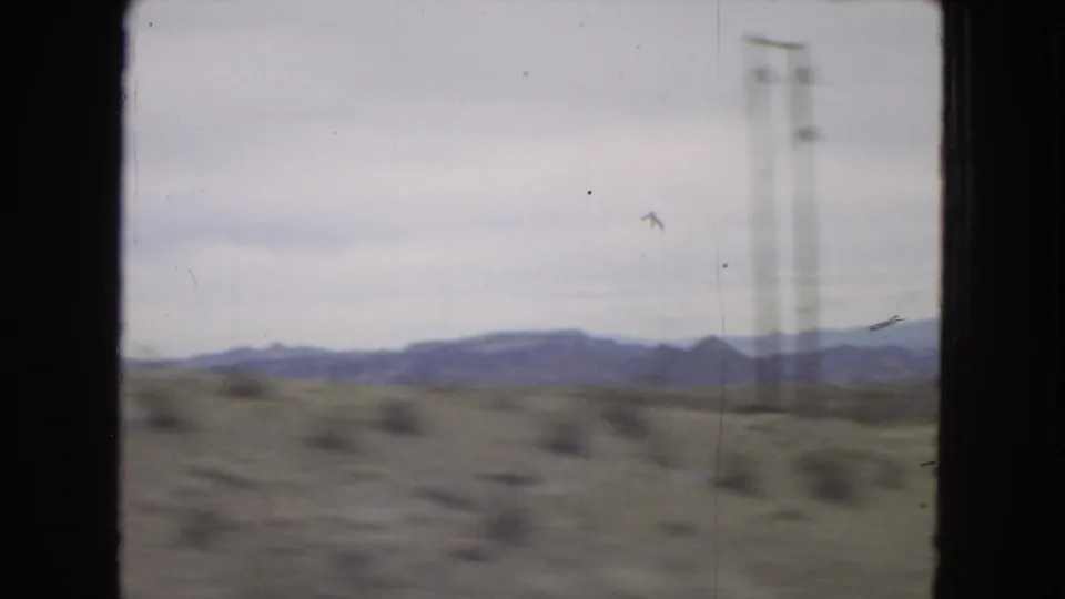 Moving-vehicle frame with blurred desert scrub in the foreground, distant blue-gray hills, cloudy sky, and a faint vertical roadside utility pole or structure.
