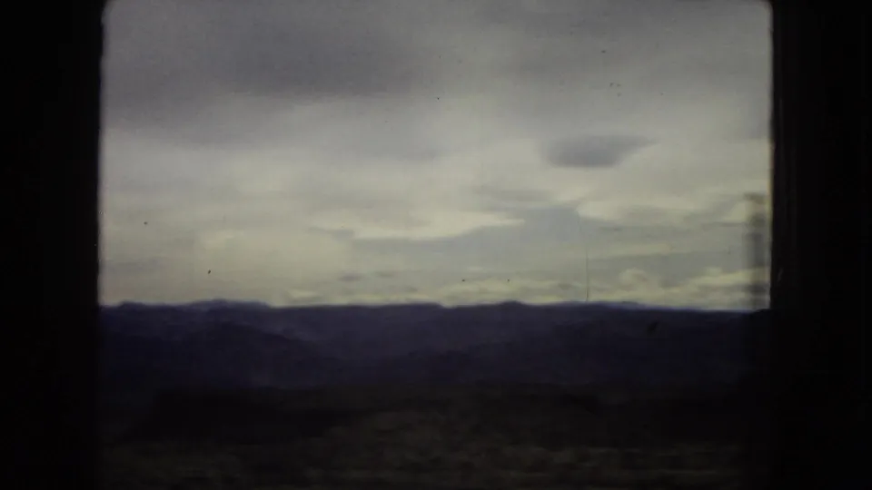 A darker 8mm frame looking across a low desert horizon toward distant mountains under a heavy cloudy sky, bordered by the vehicle window frame.