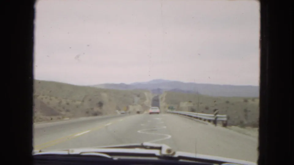 1967 archival 8mm forward-facing road view in the American Southwest, with a two-lane highway, guardrails, low hills, and a white car ahead.