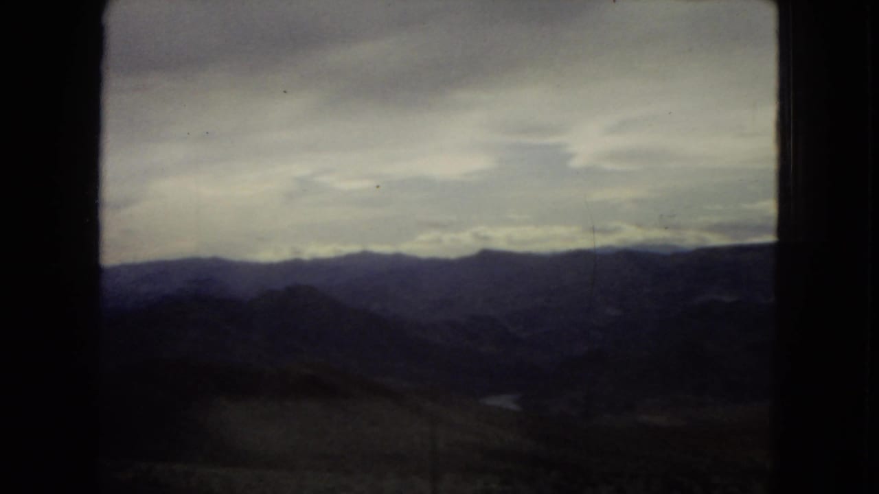 1967 8mm roadside view from inside a vehicle, showing dark window edges around arid hills, layered mountains, and a cloudy sky.
