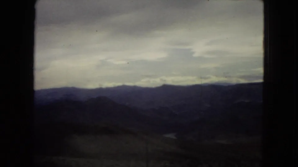 1967 8mm roadside view from inside a vehicle, showing dark window edges around arid hills, layered mountains, and a cloudy sky.
