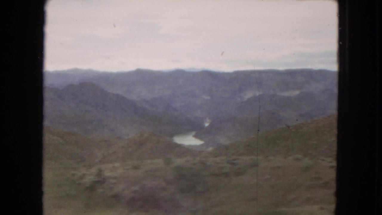 1967 Arizona home-movie frame showing scrub-covered hills, distant mountain ridges, and a winding river or reservoir in the valley.