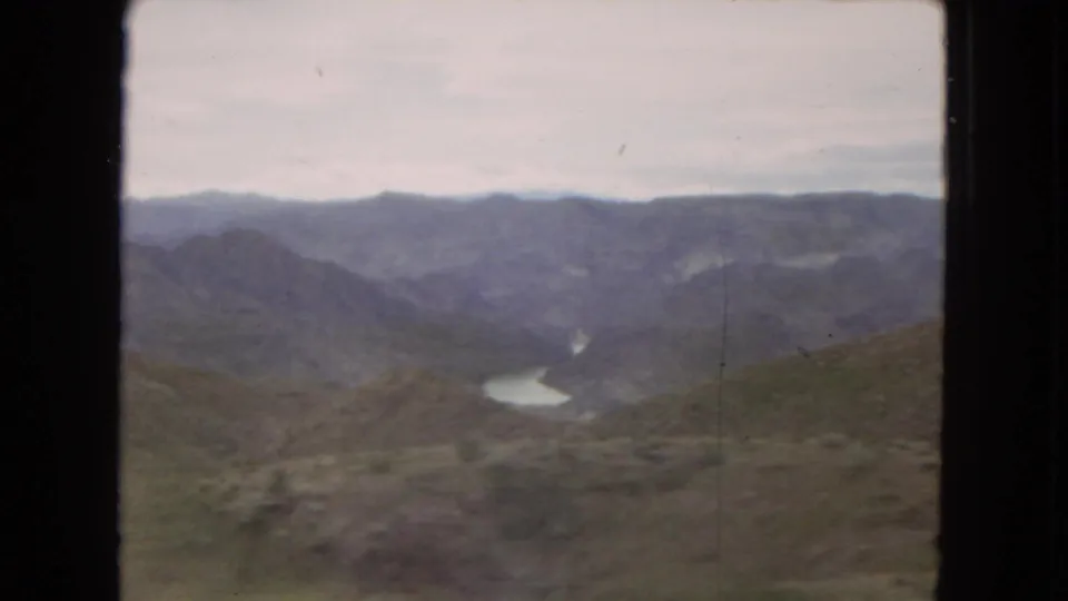 1967 Arizona home-movie frame showing scrub-covered hills, distant mountain ridges, and a winding river or reservoir in the valley.