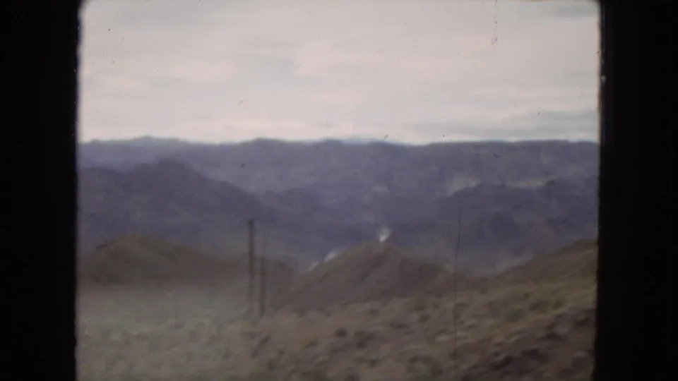 1967 8mm travel frame with hazy western mountain ridges, dry foreground hills, and a slender utility pole or powerline structure near the roadside.