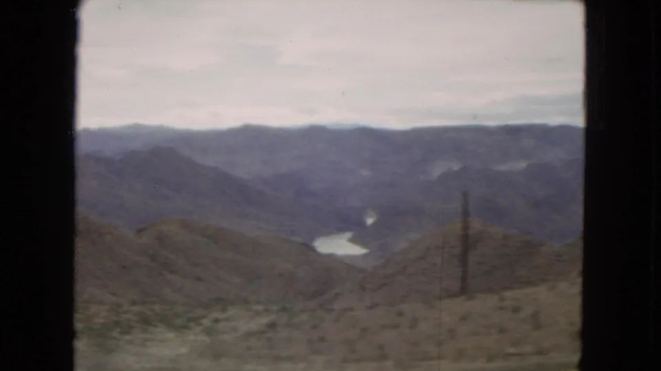 1967 Arizona roadside landscape frame showing dry hills, a distant river or reservoir, layered mountains, and a utility structure near the right side.