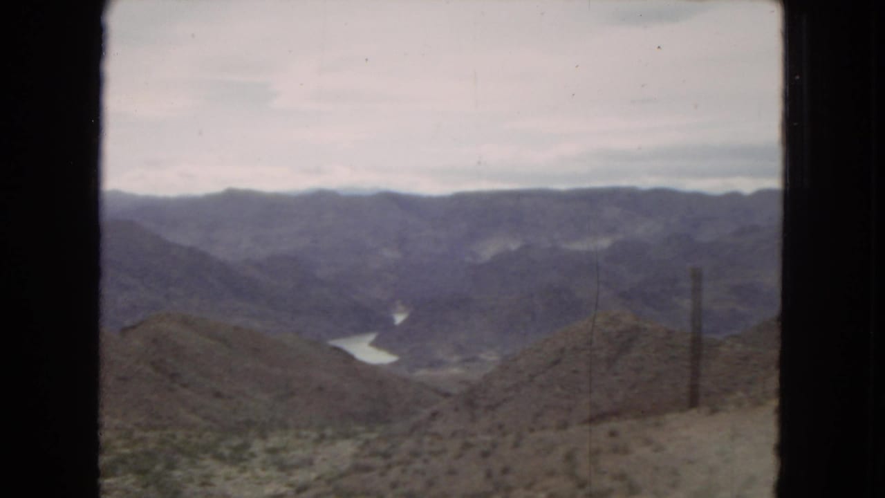 1967 Arizona home-movie view from a moving train window, showing layered desert hills, overcast sky, and a pale winding river or reservoir below.