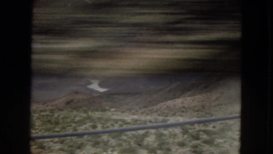 Motion-blurred 1967 Arizona train-window view with a foreground railing, scrubby hillside, layered mountains, and a winding river or reservoir in the distance.