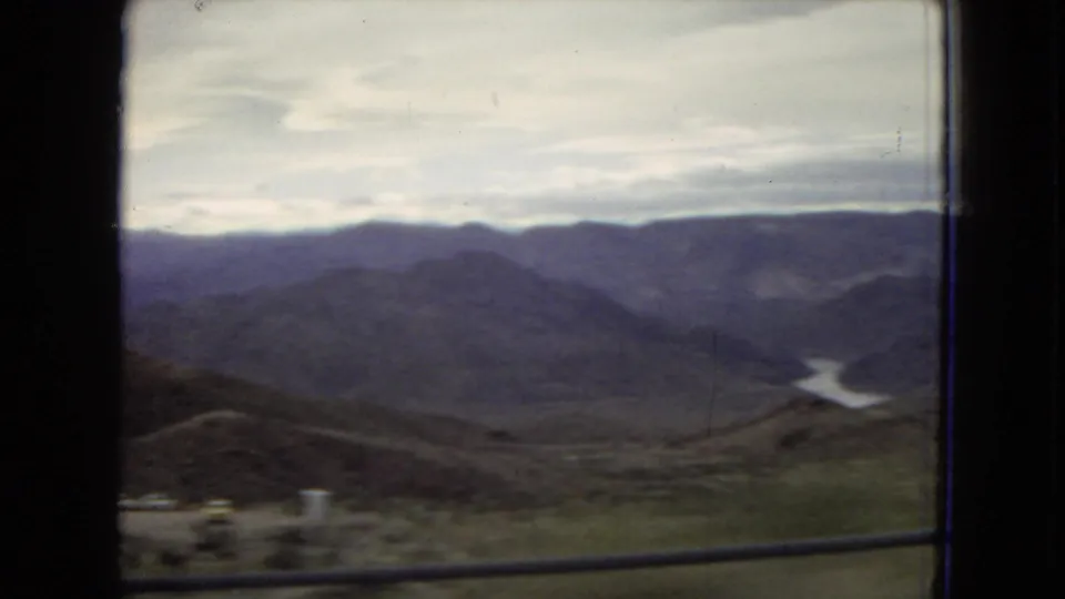 1967 Arizona home-movie frame showing a wide mountain valley from a moving train, with a foreground railing, distant water, and small structures near the lower edge.
