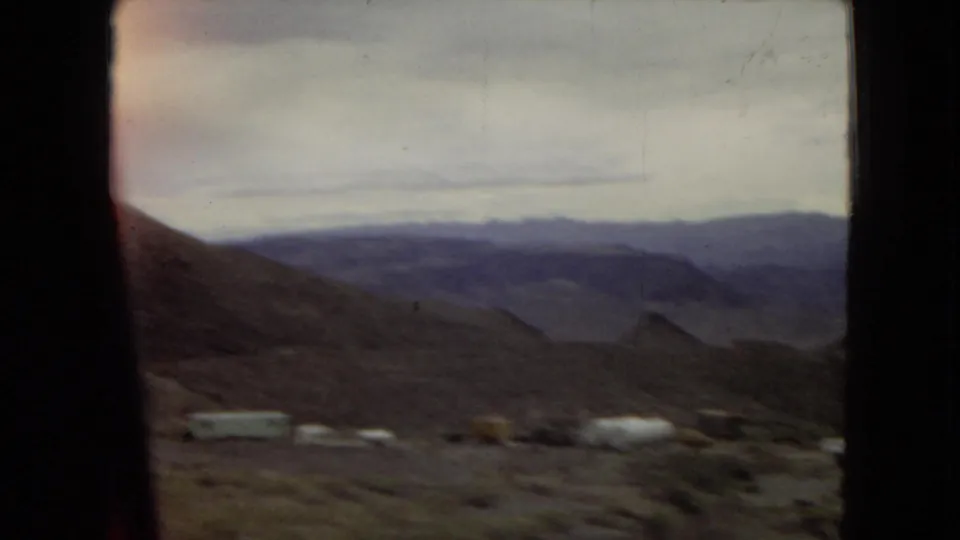 1967 Arizona train-window view across desert hills and distant mountain ridges, with several small structures or tanks visible near the bottom of the frame.