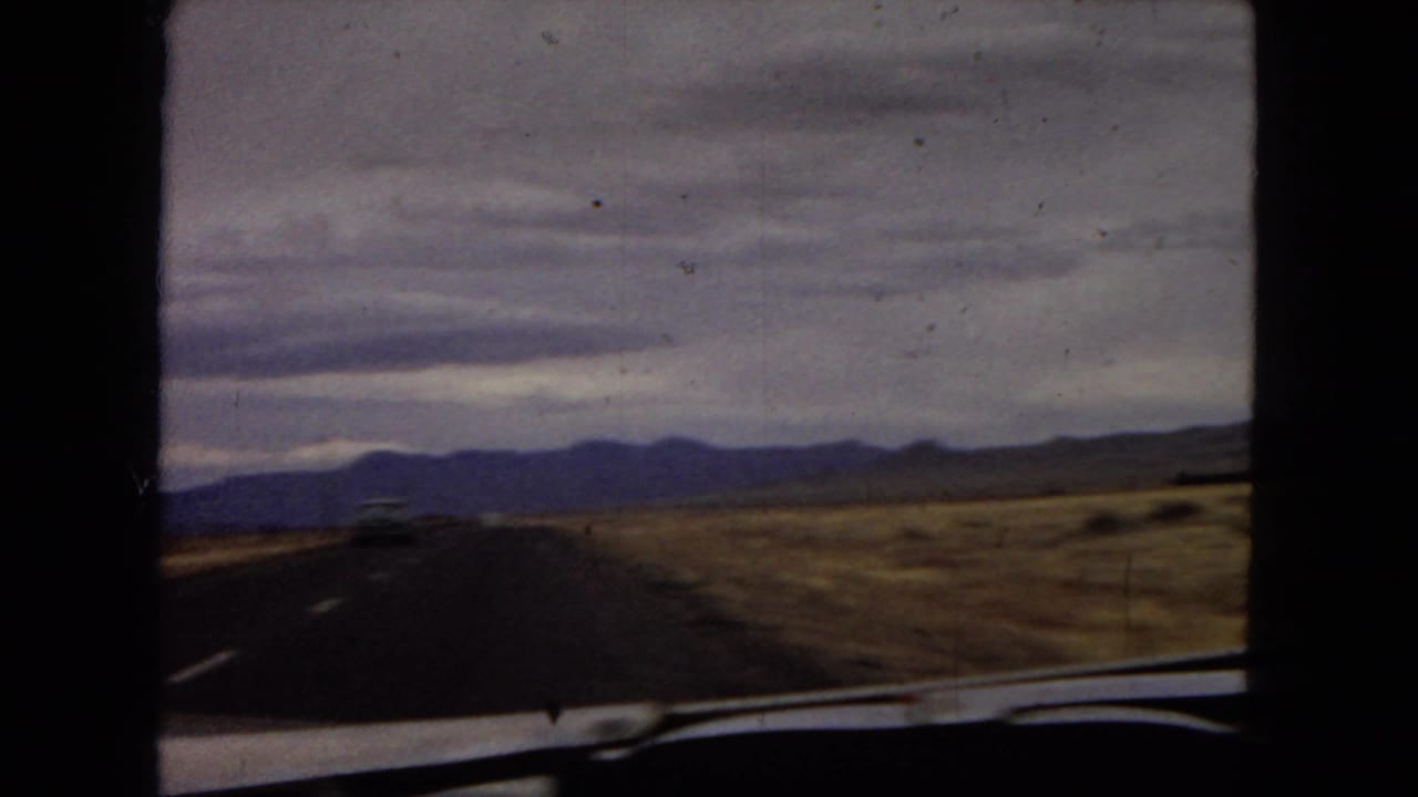 1967 home-movie view from inside a moving car on an open desert road in Arizona, with mountains ahead and the vehicle window frame crossing the foreground.