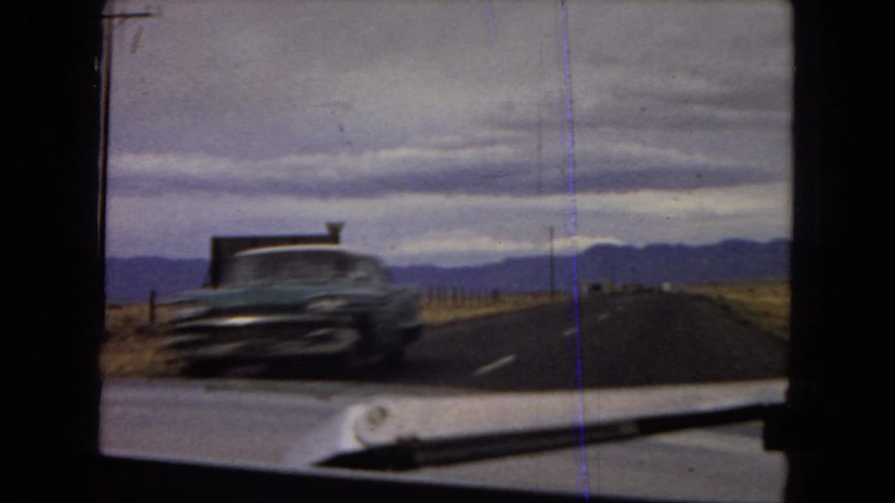 A passing mid-century car appears on the desert highway during the 1967 Arizona road-view footage, with mountains and an overcast sky in the background.