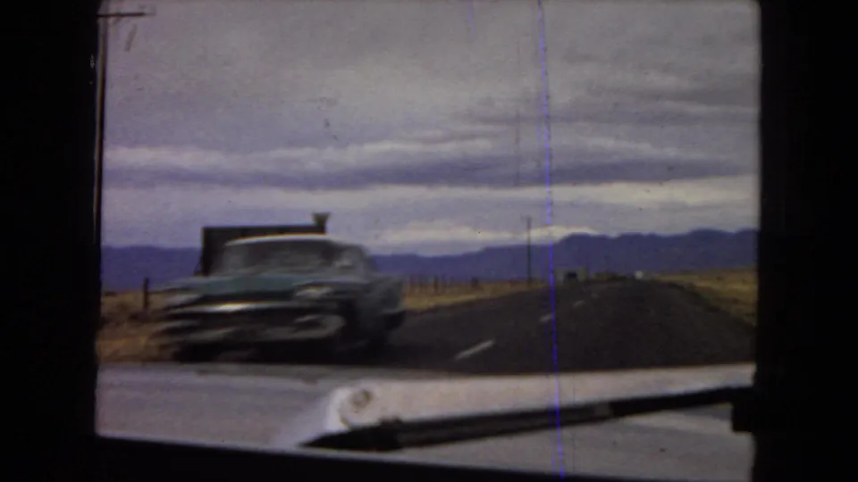 A passing mid-century car appears on the desert highway during the 1967 Arizona road-view footage, with mountains and an overcast sky in the background.
