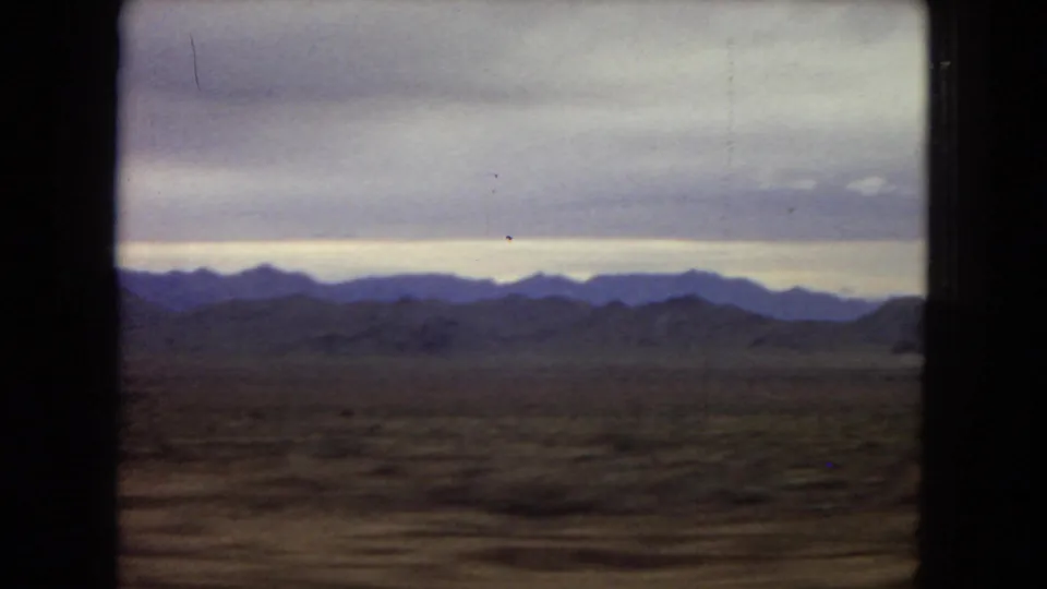 1967 Arizona home-movie frame showing a broad desert plain and distant mountains beyond the car window, with soft 8mm color and overcast light.