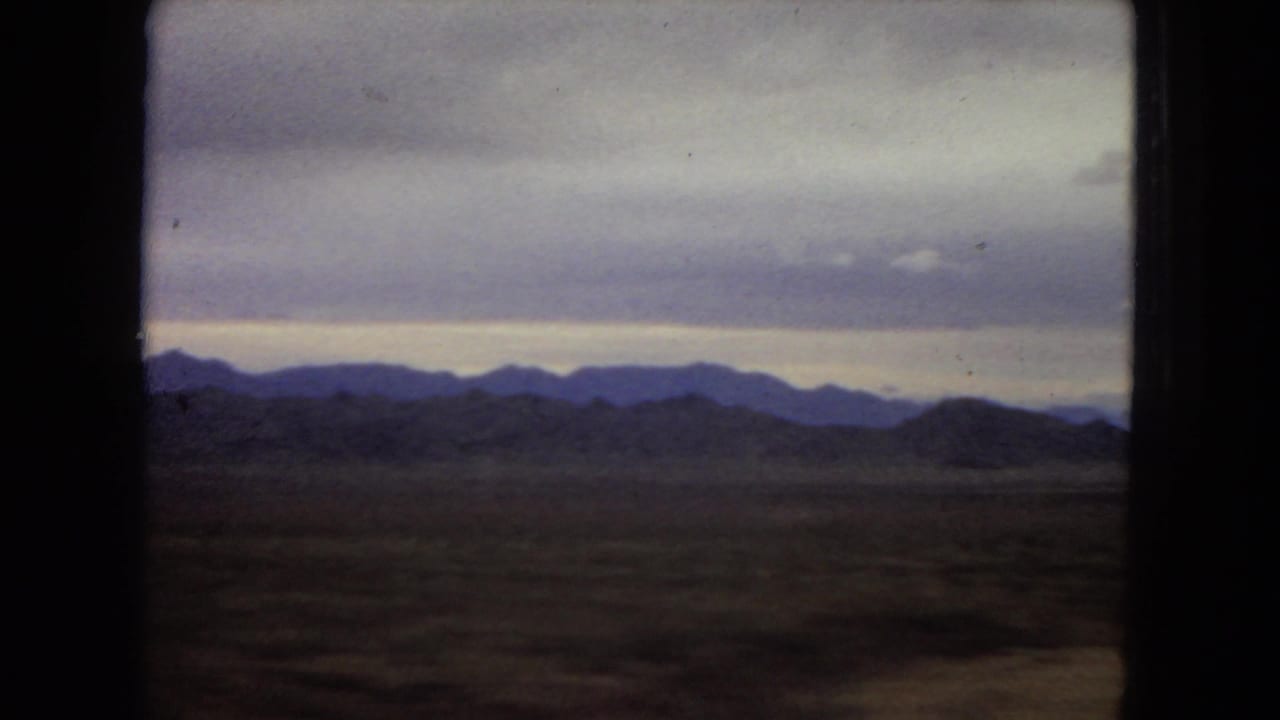 1967 Arizona 8mm home-movie frame showing layered desert mountains through a dark vehicle window, with muted clouds and a dim horizon glow.