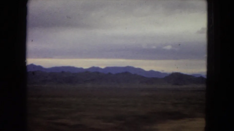 1967 Arizona 8mm home-movie frame showing layered desert mountains through a dark vehicle window, with muted clouds and a dim horizon glow.