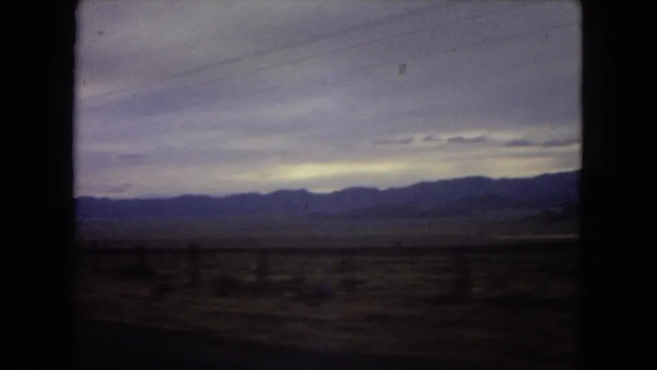 1967 Arizona travel footage frame with a distant mountain range, open desert, and purple-gray overcast sky viewed from inside a moving vehicle.
