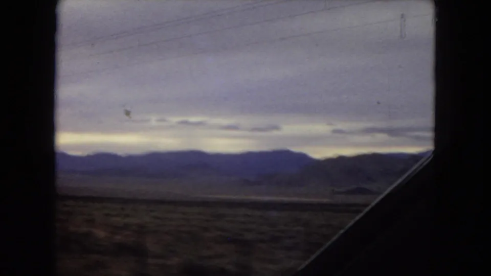1967 Arizona 8mm frame showing blurred desert foreground, layered mountains, low cloud cover, and a diagonal vehicle-window silhouette.